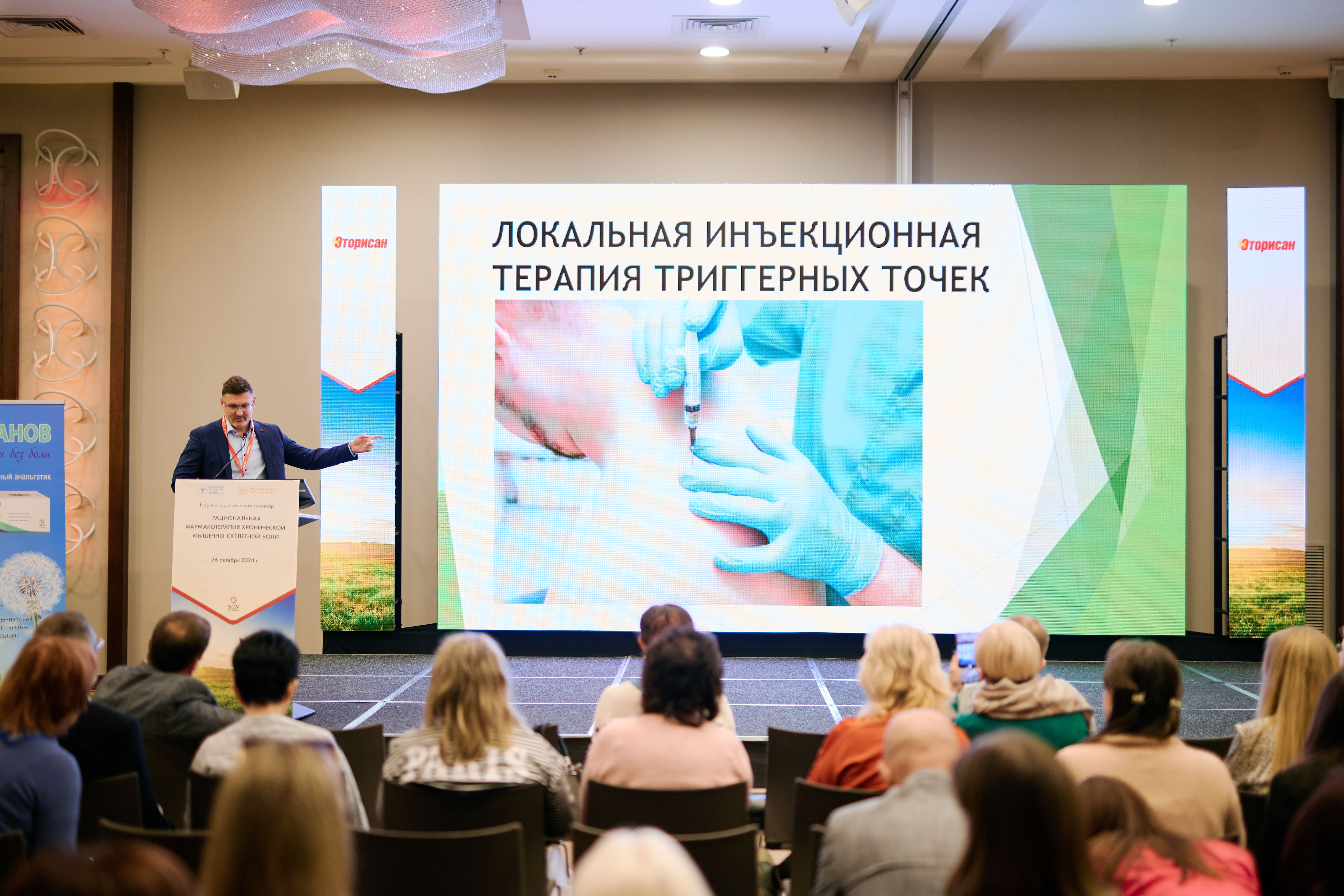 Спикер выступает перед аудиторией на фоне большого экрана с презентацией на тему "Локальная инъекционная терапия триггерных точек" в рамках конференции «Рациональная фармакотерапия хронической мышечно-скелетной боли».